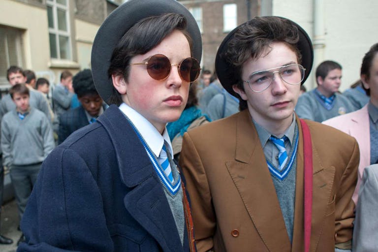 Two white teen boys in school uniforms and 80s clothing and hairstyles looking thoughtful