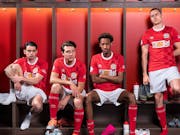 Three white and one Black young men footballers in red and white kit sitting in a locker room