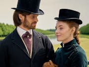 A young white man and young white woman in smart 1970s clothing and top hats stood in a large garden estate