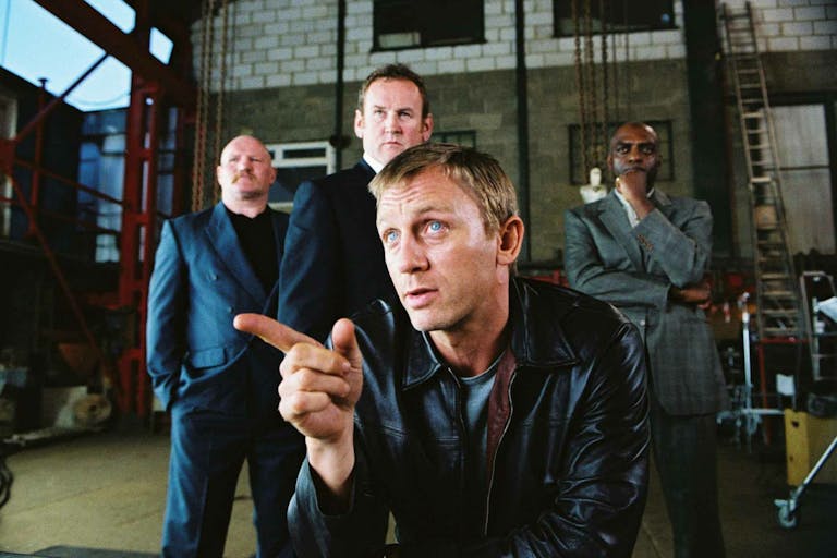 A white man in a black leather jacket sits in a warehouse, flanked by two white man and one Black man in suits