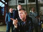 A white man in a black leather jacket sits in a warehouse, flanked by two white man and one Black man in suits