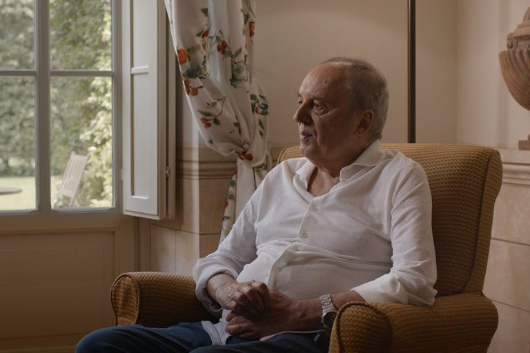 An older white man in a white shirt sits in an armchair by a bright window