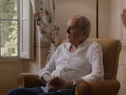 An older white man in a white shirt sits in an armchair by a bright window