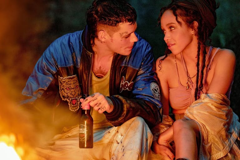 A young white man with tattoos holding a beer looks admiringly at a young mixed heritage Black woman with dreads as they sit by a fire