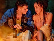 A young white man with tattoos holding a beer looks admiringly at a young mixed heritage Black woman with dreads as they sit by a fire