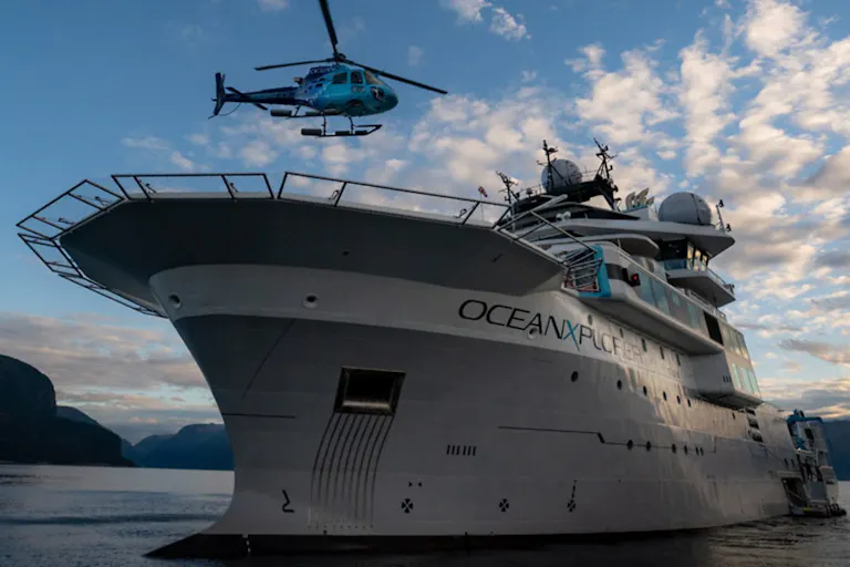 A large white boat with OceanXplorer written on the side in the sea with a helicopter flying overhead