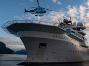 A large white boat with OceanXplorer written on the side in the sea with a helicopter flying overhead
