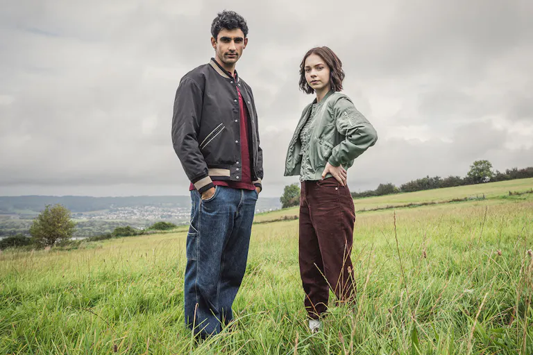 A young brown man and young white girl stand in a green field looking to camera