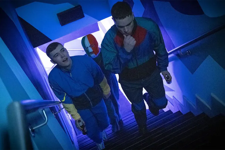 Three young white men walking up purple lit staircase, wearing tracksuits, one wearing an Irish tricolour balaclava