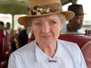 An old white woman in a white blouse and a straw hat with flowers on, sitting on a bus