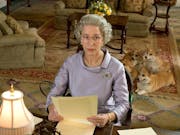 An old white woman with short grey hair in regal dress suit sitting at a desk with four corgis in the background