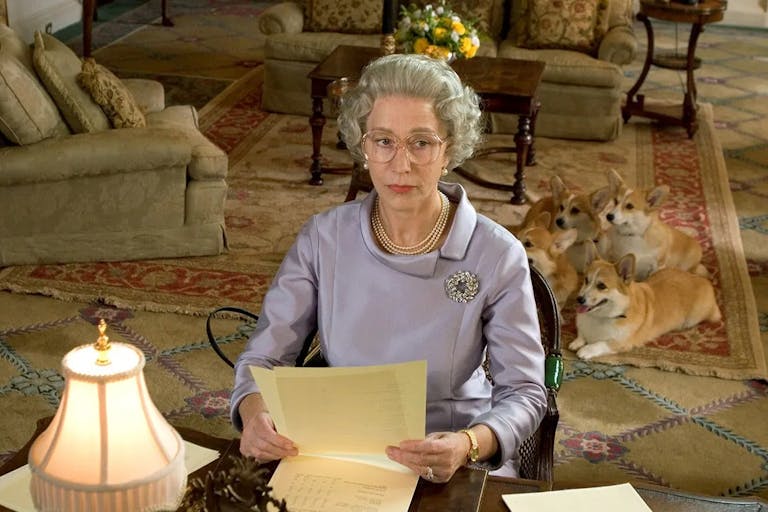 An old white woman with short grey hair in regal dress suit sitting at a desk with four corgis in the background