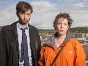 A white man in a suit and white woman in an orange raincoat looking serious, with a coastal cliff behind them