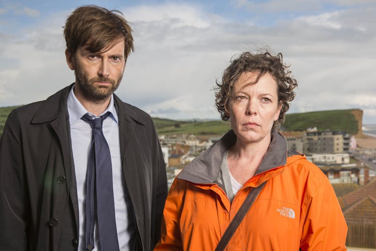 A white man in a suit and white woman in an orange raincoat looking serious, with a coastal cliff behind them