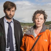 A white man in a suit and white woman in an orange raincoat looking serious, with a coastal cliff behind them