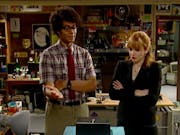 A Black man in a checked shirt and tie and a white ginger woman in a suit stand over a small metal box on a messy basement office