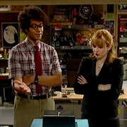 A Black man in a checked shirt and tie and a white ginger woman in a suit stand over a small metal box on a messy basement office