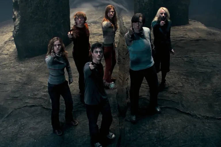 A group of young white people standing in a dark stone place, looking dishevelled pointing wands up in the air