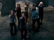 A group of young white people standing in a dark stone place, looking dishevelled pointing wands up in the air