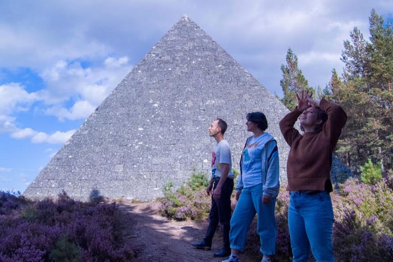 Three people stand in the bright outdoors by a medium sized grey stone pyramid