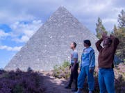 Three people stand in the bright outdoors by a medium sized grey stone pyramid