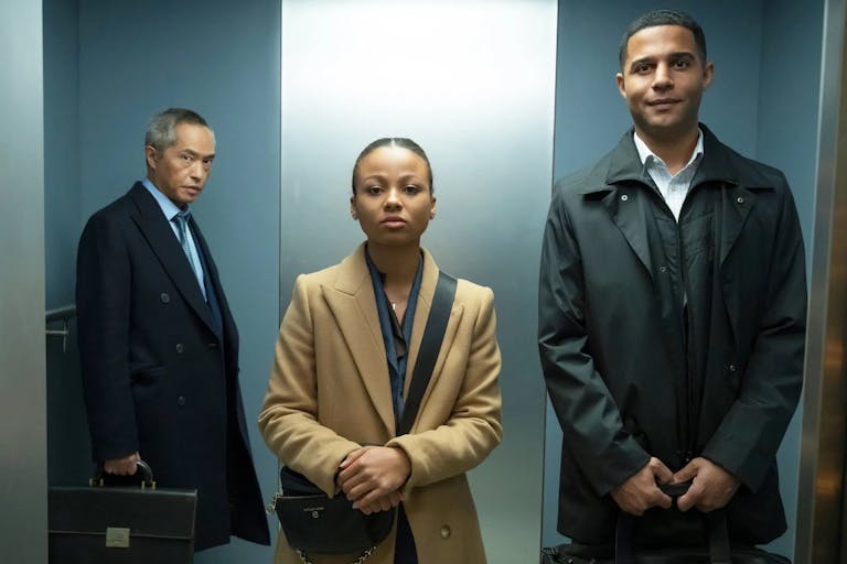 A young Black woman and man in suits stand in a lift with an older American Chinese man in the background