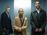 A young Black woman and man in suits stand in a lift with an older American Chinese man in the background