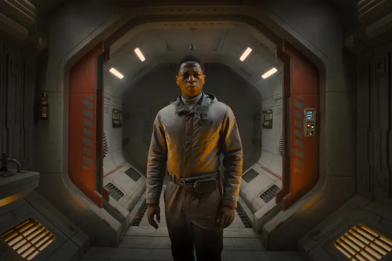 A young Black man in in brown overalls stands in a cinematic spaceship entrance looking shocked