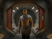 A young Black man in in brown overalls stands in a cinematic spaceship entrance looking shocked