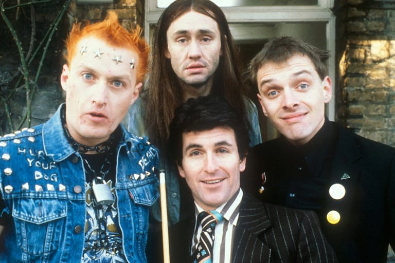 Four young white men, one with spiky punk hair and clothing, one in a suit and tie, one in all black clothing and one with long black hair