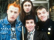 Four young white men, one with spiky punk hair and clothing, one in a suit and tie, one in all black clothing and one with long black hair