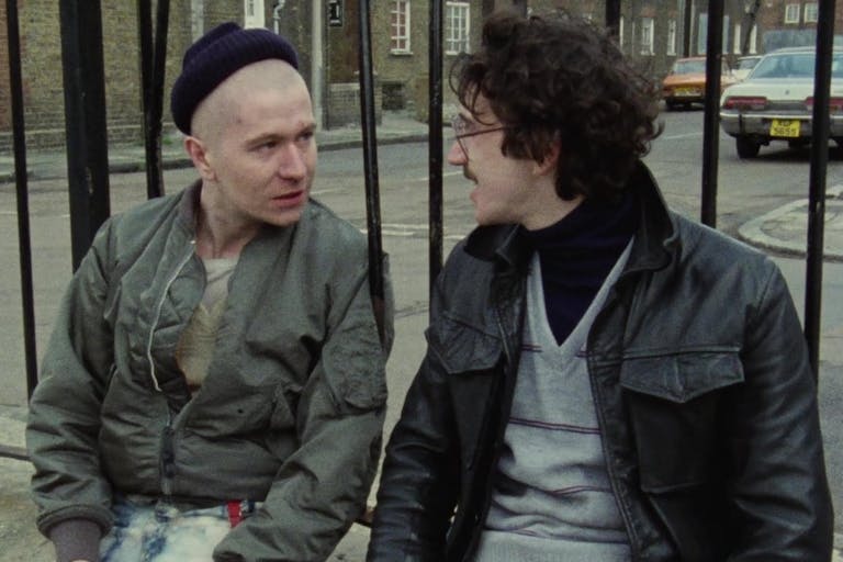 A bald, young white thuggish man in Harrington jacket sits next to a young white man in a woollen vest against black railings