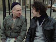 A bald, young white thuggish man in Harrington jacket sits next to a young white man in a woollen vest against black railings