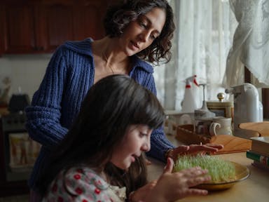 A young Iranian woman with a young Iranian girl at a kitchen counter tending to a small dish of growing plants