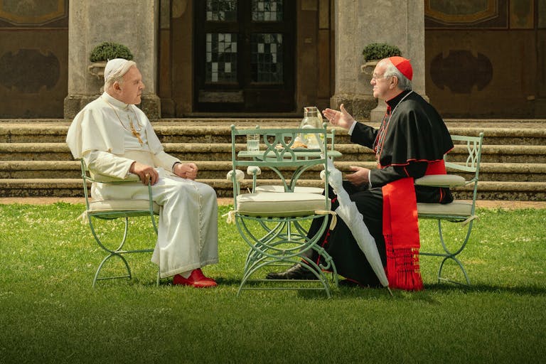 Two old white men in religious catholic pope attire, one in white, one in black and red, sitting on garden furniture on a manicured lawn