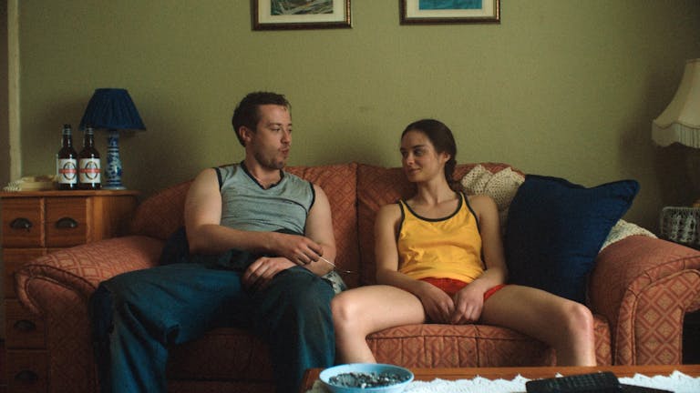 A young white man and a teenage girl sit on a sofa looking at each other, bottles of beer on a table next to them