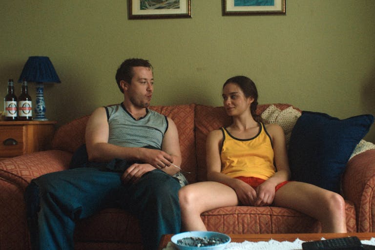 A young white man and a teenage girl sit on a sofa looking at each other, bottles of beer on a table next to them