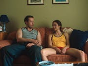 A young white man and a teenage girl sit on a sofa looking at each other, bottles of beer on a table next to them