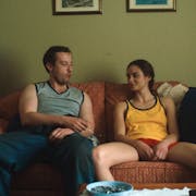 A young white man and a teenage girl sit on a sofa looking at each other, bottles of beer on a table next to them