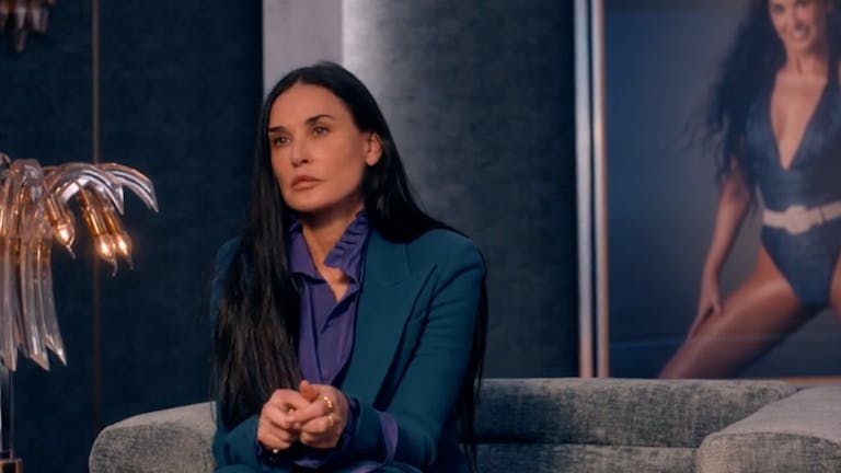 A white woman in a stylish suit sitting on a designer sofa with a large print of her younger self in the background