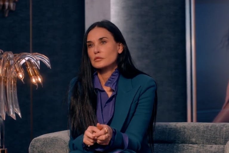 A white woman in a stylish suit sitting on a designer sofa with a large print of her younger self in the background