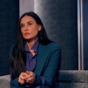 A white woman in a stylish suit sitting on a designer sofa with a large print of her younger self in the background