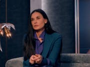 A white woman in a stylish suit sitting on a designer sofa with a large print of her younger self in the background
