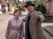 A middle aged white woman in a silk dressing gown and middle aged white man in a suit, trench coat and hat on a suburban street