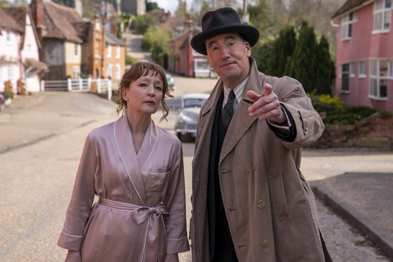 A middle aged white woman in a silk dressing gown and middle aged white man in a suit, trench coat and hat on a suburban street