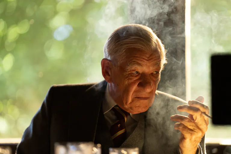An old white man sitting at a desk smoking a cigarette looking menacing
