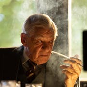An old white man sitting at a desk smoking a cigarette looking menacing