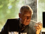 An old white man sitting at a desk smoking a cigarette looking menacing