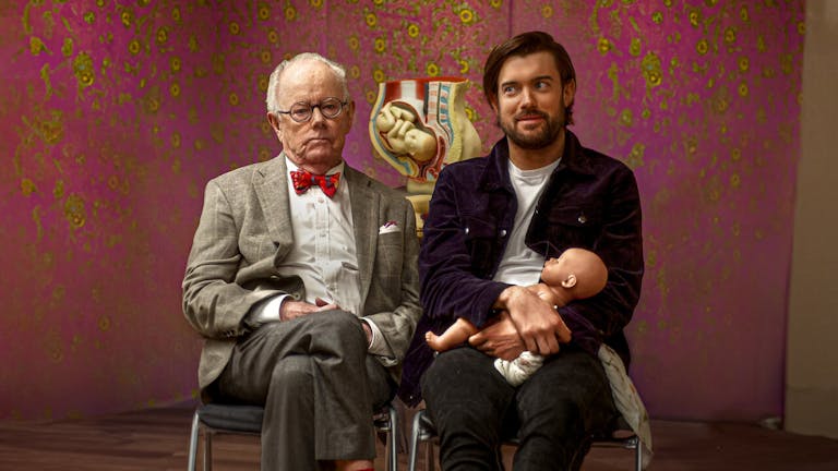 An older grey white man in a suit and glasses sitting next to a white man holding a doll with a cross section model of a pregnant belly behind them