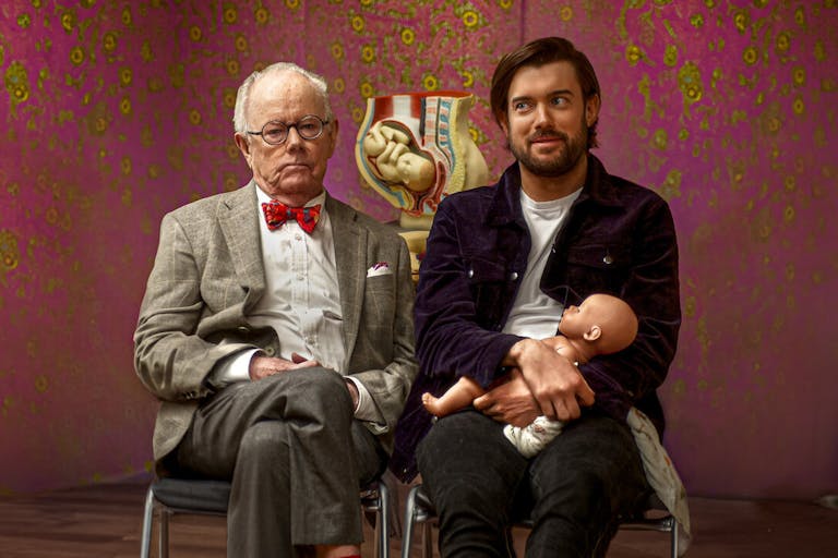 An older grey white man in a suit and glasses sitting next to a white man holding a doll with a cross section model of a pregnant belly behind them